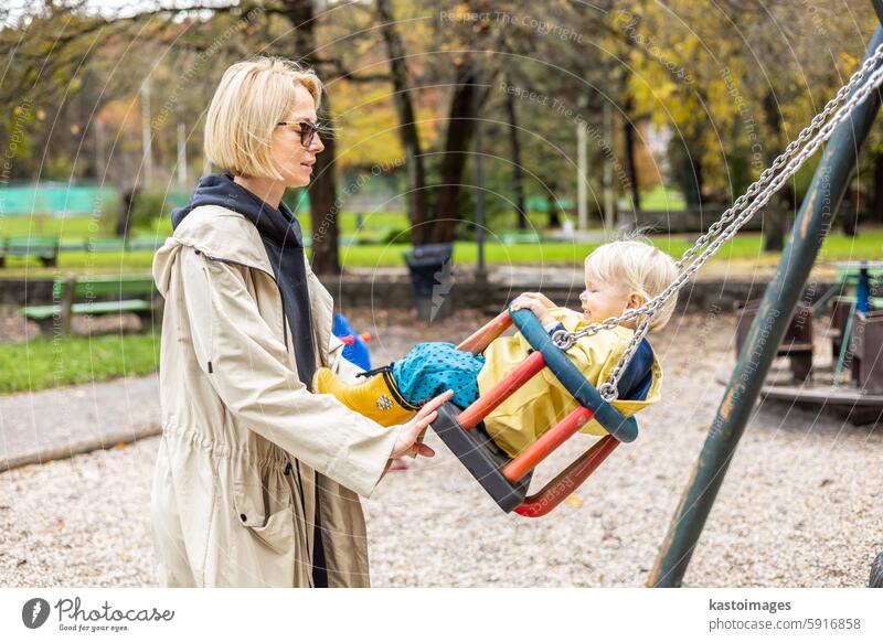 Mutter schiebt ihr Kleinkind mit gelben Regenstiefeln und Umhang auf der Schaukel auf dem Spielplatz im Freien an einem kalten, regnerischen, bedeckten Herbsttag in Ljubljana, Slowenien