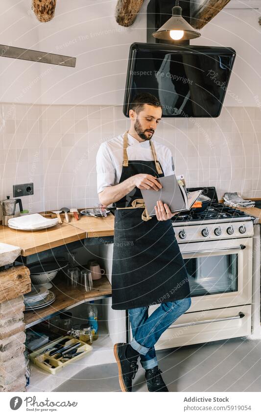 Privatkoch liest Rezept in der Küche und schaut nach unten Küchenchef privat männlich professionell Essen zubereiten Buch herabsehend Schürze Herd rustikal