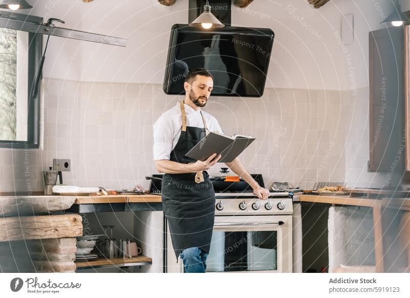 Privatkoch in moderner Küche bei der Überprüfung von Rezepten Küchenchef Buch Schürze männlich Essen zubereiten Lebensmittel professionell kulinarisch