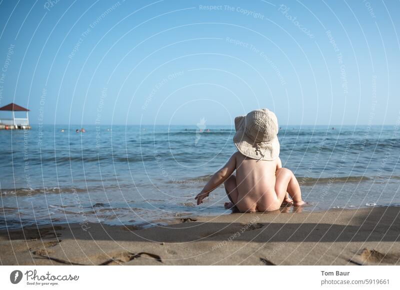 Baby mit Hut am Strand blickt ins Meer Kind niedlich klein schön süß Kaukasier weiß weich unschuldig Ferne Wellen Sandstrand Sonnenhut Blauer Himmel