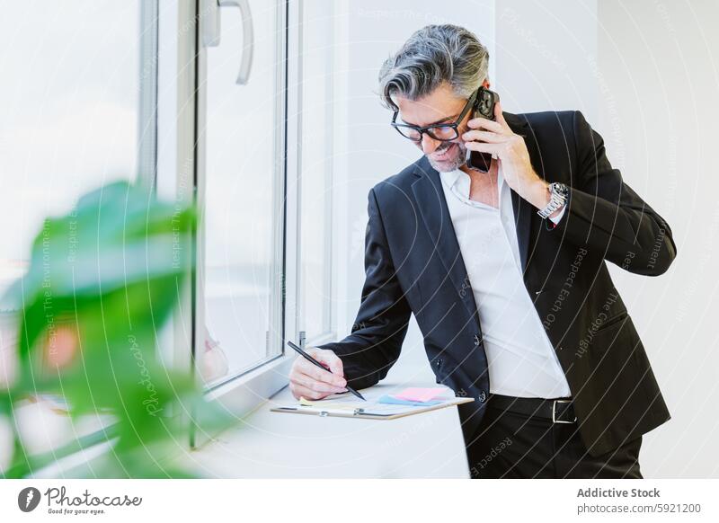 Ein konzentrierter Manager, der sein Arbeitspensum mit einem Telefon zwischen Schulter und Ohr und einem Stift in der Hand vor einem hellen Fenster bewältigt