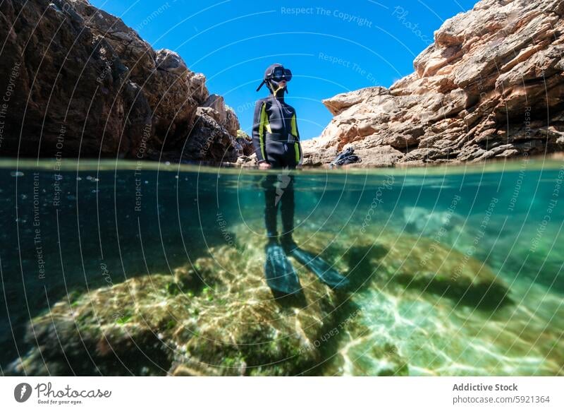 Unbekannter Jugendlicher in Neoprenanzug und Schnorchel im klaren Wasser mit felsigen Klippen von Menorca, Spanien, geteilter Blick über und unter der Wasseroberfläche