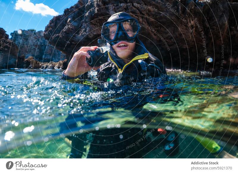 Porträt eines anonymen männlichen Teenager-Tauchers, der einen Neoprenanzug und eine Maske trägt und in die Kamera schaut, während er in den Sommerferien im Meer gegen Felsformationen schwimmt