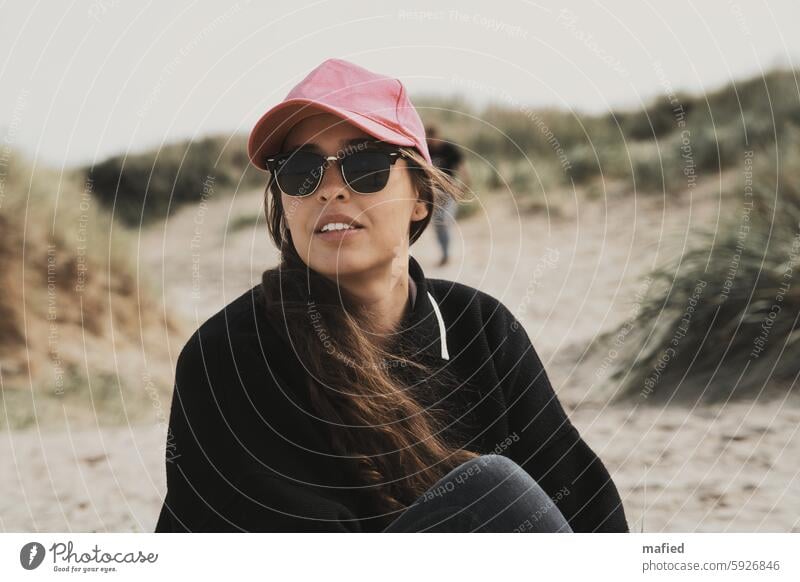 Junge Frau mit Sonnenbrille und Baseball Cap sitzt am Strand jung Dünen Blick in die Ferne Porträt Erwachsene Außenaufnahme feminin 18-30 Jahre Sommertag