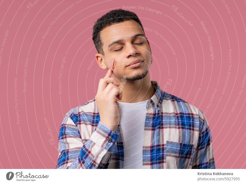 Junger Mann mit Rosenquarzsteinrolle auf rosa Hintergrund. Selbstpflege im Gesicht Erwachsener Anti-Aging attraktiv schön Schönheit Körperpflege Pflege