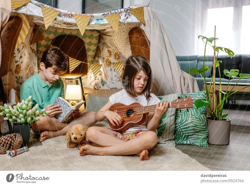 Kleines Mädchen spielt Ukulele, während ein Junge ein Buch auf einem handgefertigten Tipi im Wohnzimmer liest Spielen Gitarre lesen Kinder habend Spaß