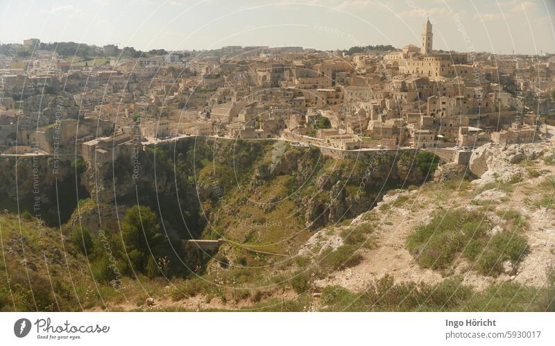 Blick auf die Schlucht vor der Stadt und die Altstadt von Matera (Süditalien) im Mittagslicht. Man siegt uralte Häuser aus Felssteinen und in den Fels gehauene Wohnhöhlen. Im Hintergrund eine Kirche.