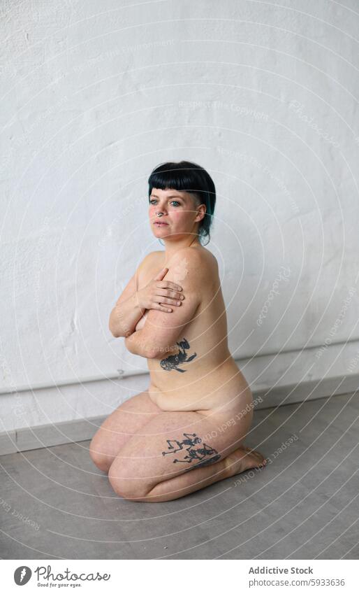 Frau mit Tattoos, die vor einem schlichten Hintergrund sitzt Porträt Ausdruck neutral Sitzen im Innenbereich Schwarzes Haar Bobschnitt Haut Tattoo-Kunst Pose