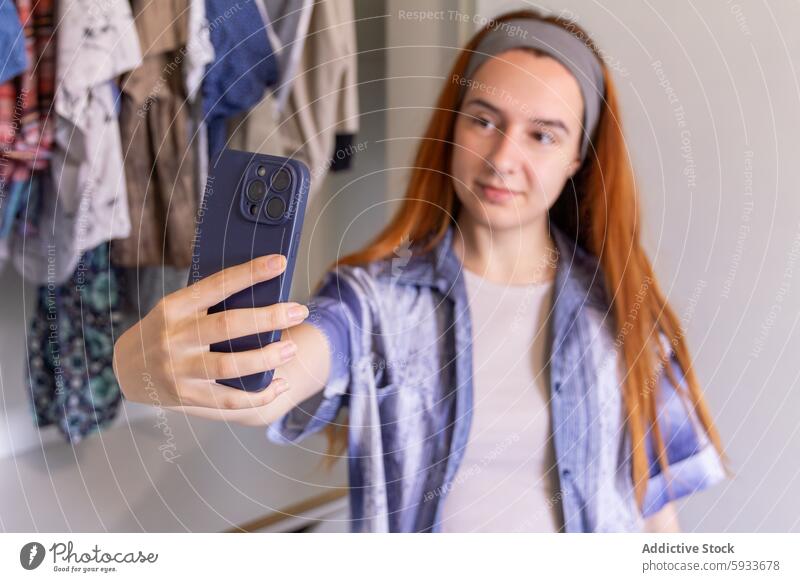 Junge Frau, die ihren Kleiderschrank sortiert und Kleidung online verkauft Verkauf Smartphone Selfie Fotokamera Hemd rote Haare Mode Entrümpelung Lager