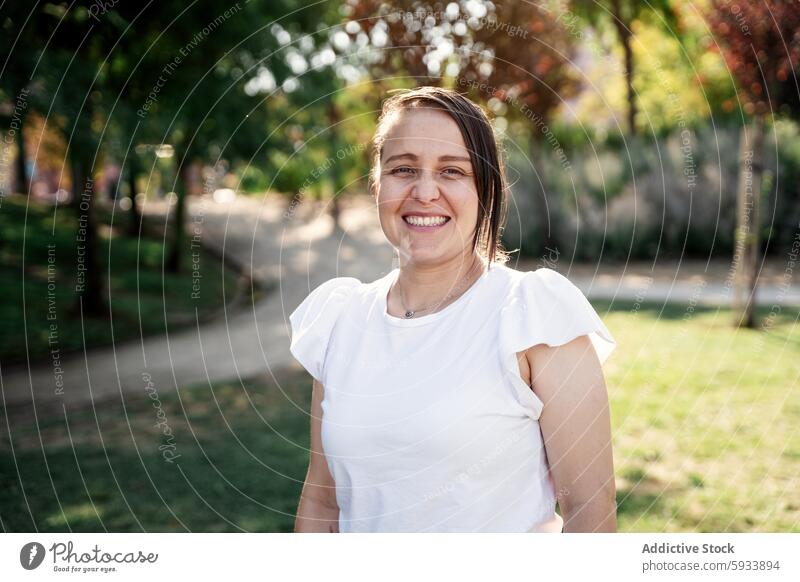 Lässige kolumbianische Frau lächelnd in sonnenbeschienenen Park mit unscharfen Baum Hintergrund Lächeln lässig weiß Bluse Glück im Freien Natur Grün Porträt