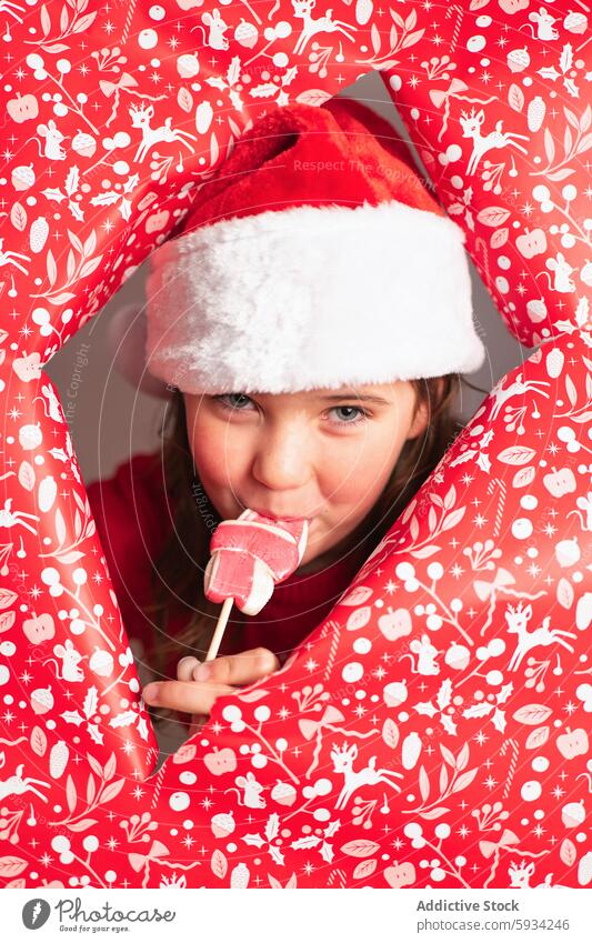 Junges Mädchen mit Weihnachtsmannmütze genießt einen Lutscher in weihnachtlicher Umgebung Weihnachtsmütze Weihnachten Lollipop Spaß spielerisch rot Thema Papier