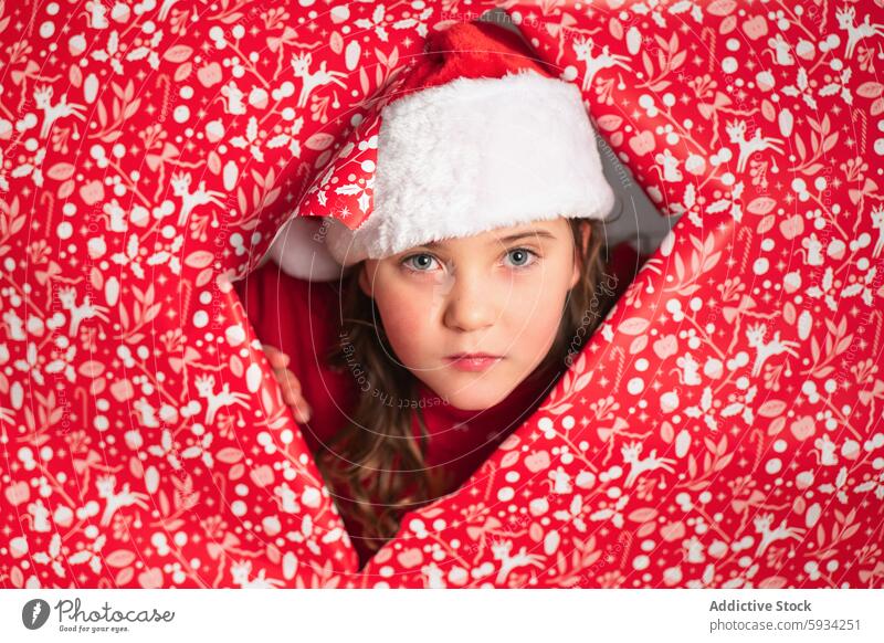 Junges Mädchen mit Weihnachtsmannmütze lugt aus roter Festtagspackung hervor Weihnachten Weihnachtsmütze festlich Muster Feiertag umhüllen schauen Sie Kind