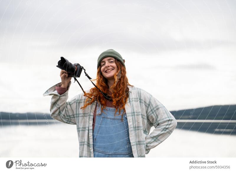 Lächelnde Frau, die beim Camping an einem See Fotos macht Fotokamera Fotografie im Freien Natur heiter Rotschopf Beanie Latzhose entspannt Hobby Freizeit Freude