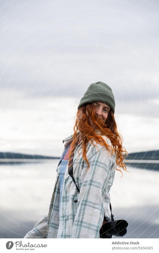 Junge Frau genießt einen Campingausflug am See im Freien Beanie Lächeln Natur Fotokamera Gelassenheit wolkig rote Haare kariertes Hemd Fotografie Freizeit