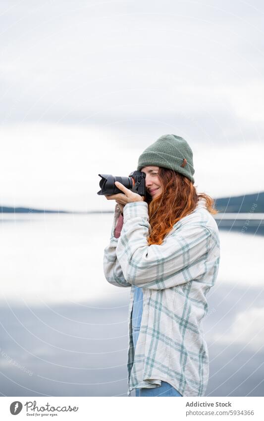 Rothaarige Frau fängt die Natur mit der Kamera am See ein Rotschopf Fotokamera Fotografie Beanie Plaid Hemd im Freien malerisch friedlich Wasser Hobby Freizeit