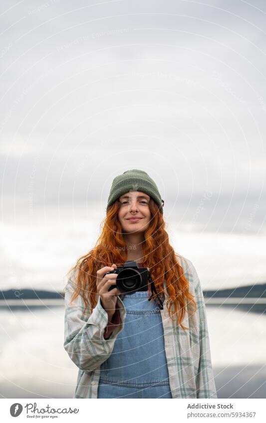 Rothaarige Frau mit Kamera bei einem Outdoor-Abenteuer Fotokamera rote Haare Beanie Jeansstoff Latzhose See Himmel wolkig im Freien Gelassenheit Natur einfangen