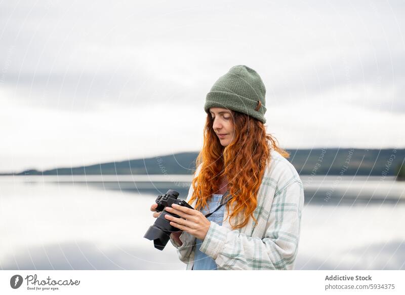 Junge Frau mit Kamera an einem ruhigen See Natur im Freien Rotschopf Beanie erkundend Ruhe Landschaft malerisch Ansicht Wasser Reflexion & Spiegelung Himmel