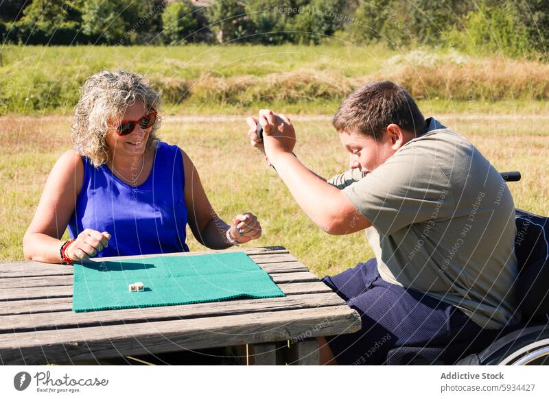 Junge im Rollstuhl spielt Würfelspiel mit Frau im Freien zerebrale Kinderlähmung Spiel Lächeln Tisch Feld Himmel grün blau Interaktion Gesundheit Behinderung