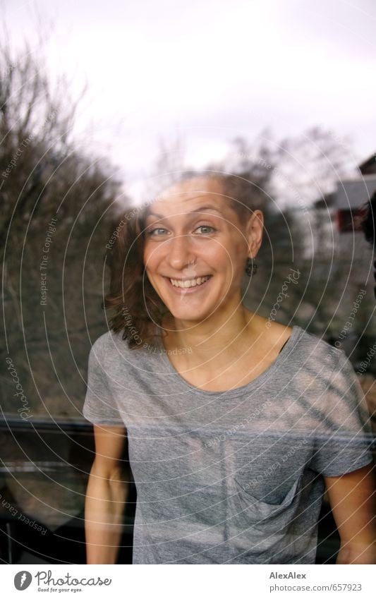 Portrait einer lachenden jungen Frau hinter einer Fensterscheibe Junge Frau Jugendliche Haare & Frisuren Gesicht 18-30 Jahre Erwachsene T-Shirt brünett